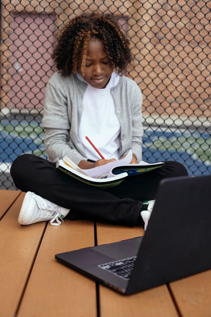 Attentive ethnic schoolchild taking notes on paper while sitting with crossed legs near netbook and doing homework